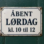 Emaljeskilt på en grøn dør med teksten Åbent lørdag kl. 10 til 12.