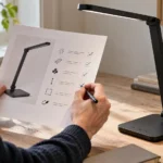 En person holder et ark med afkrydsninger ved siden af en sort skrivebordslampe og et USB-C-kabel på et træbord.